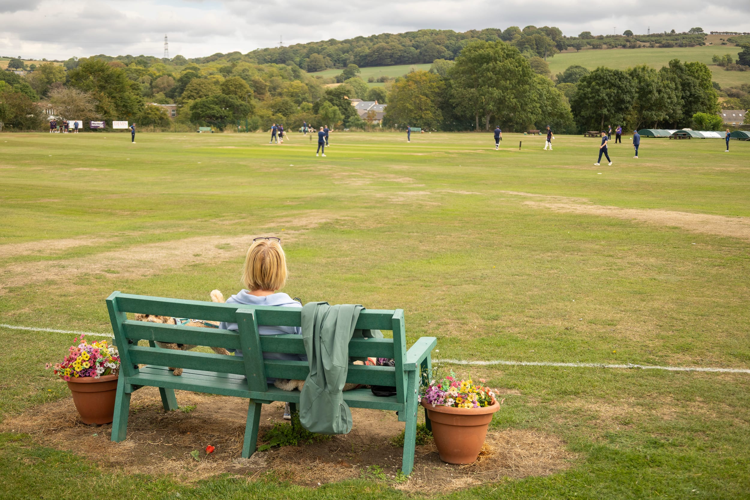 A spectator sitting on a bench watching a match on the cricket field at Lanchester, with the valley beyond.