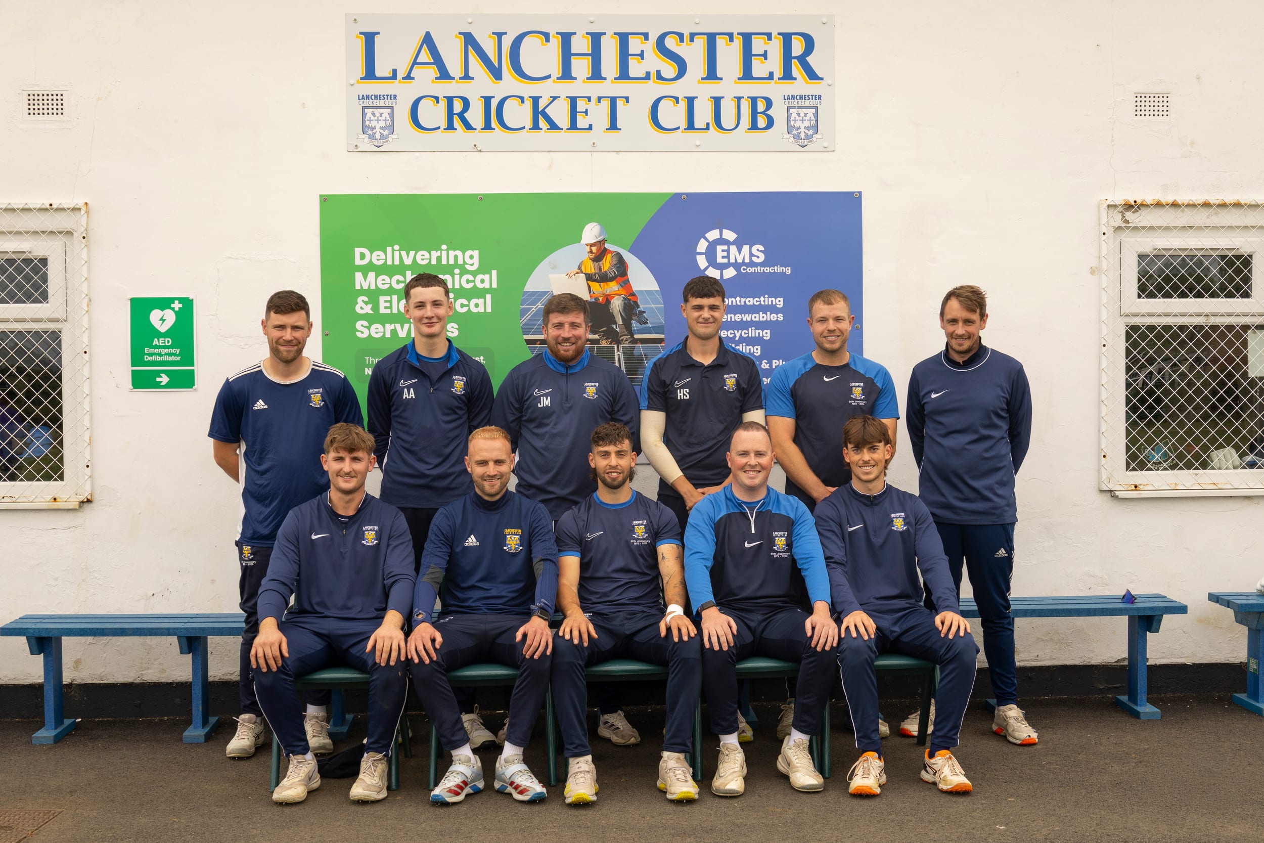 A team photograph of Lanchester Cricket Club players in blue kit seated and standing in front of the clubhouse.