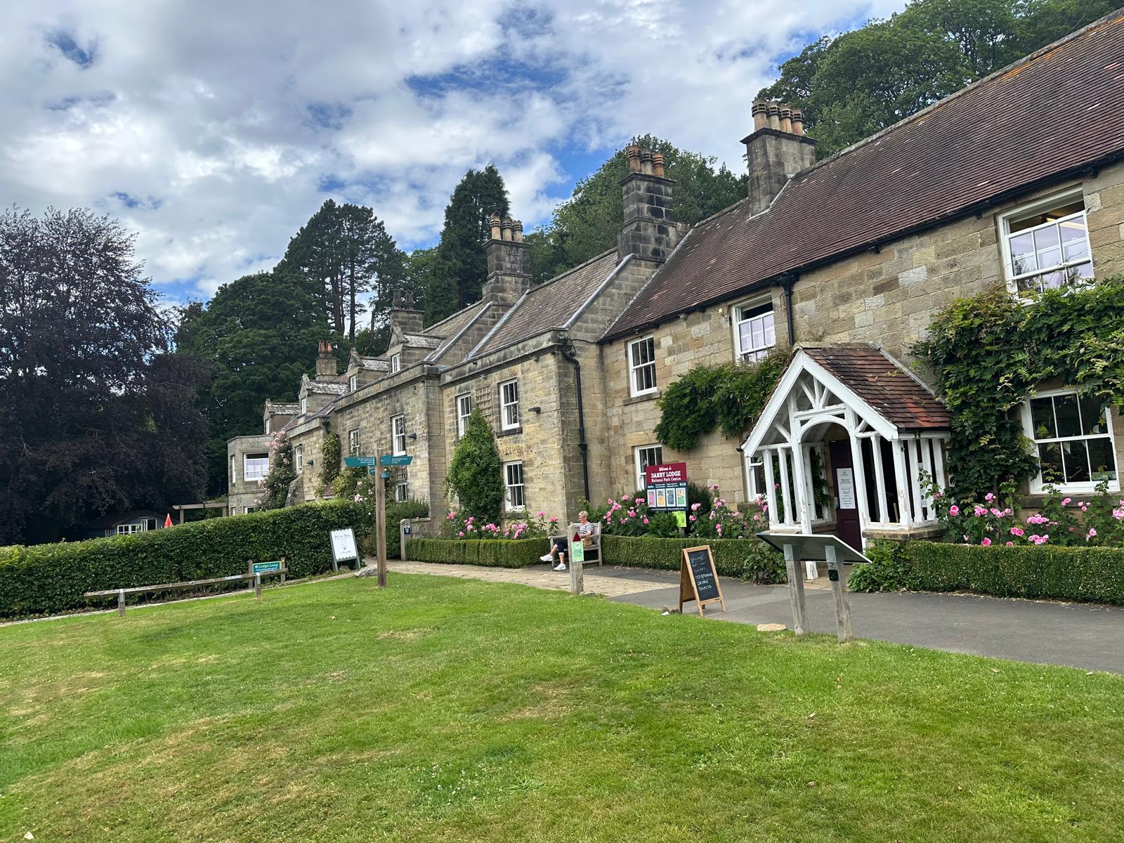 The long stone frontage of Danby Lodge National Park Centre, with climbing roses, a white porch and a lawn in front