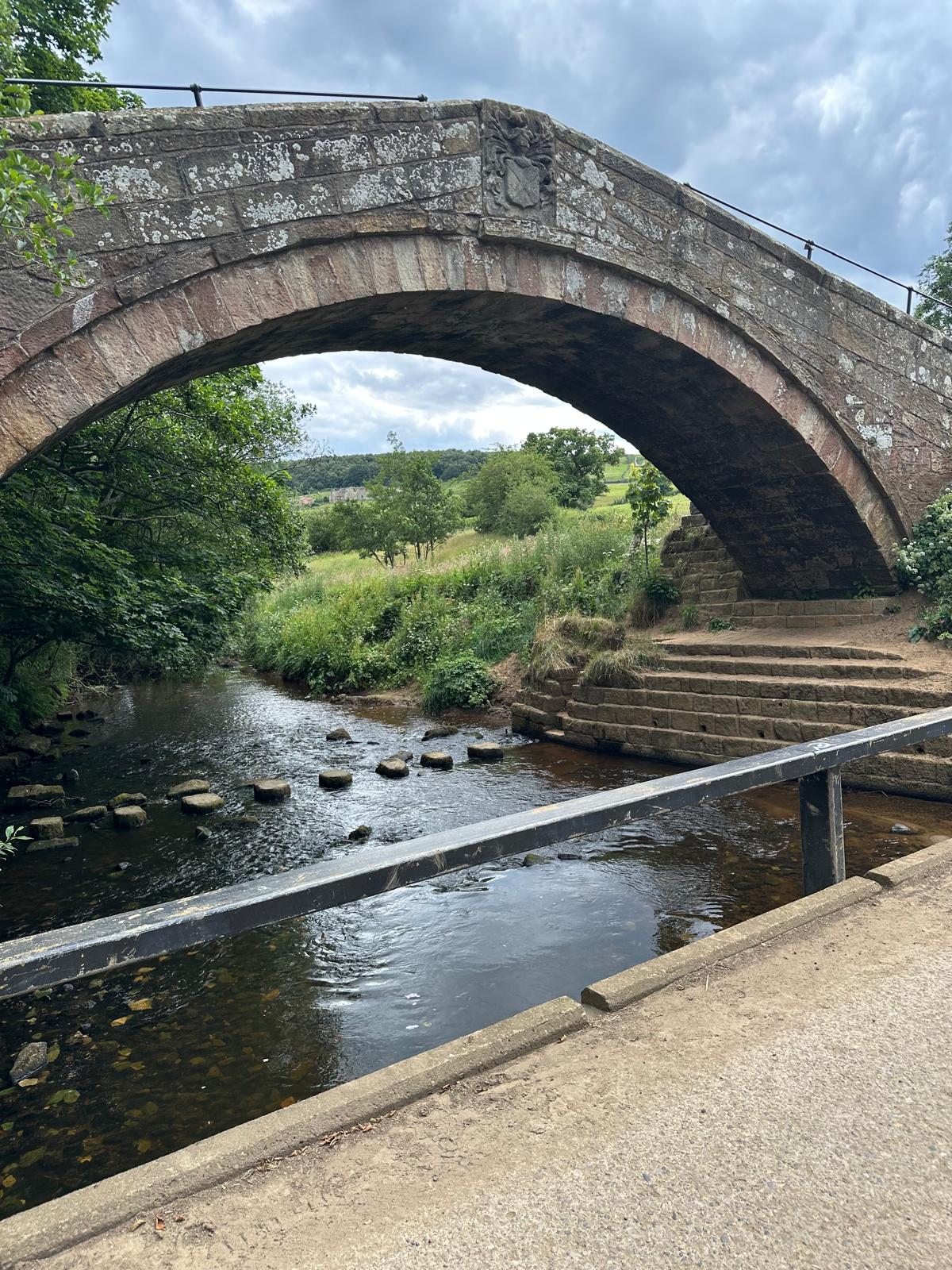 A hump-backed medieval stone bridge over a shallow river, with stepping stones crossing the water beneath the arch