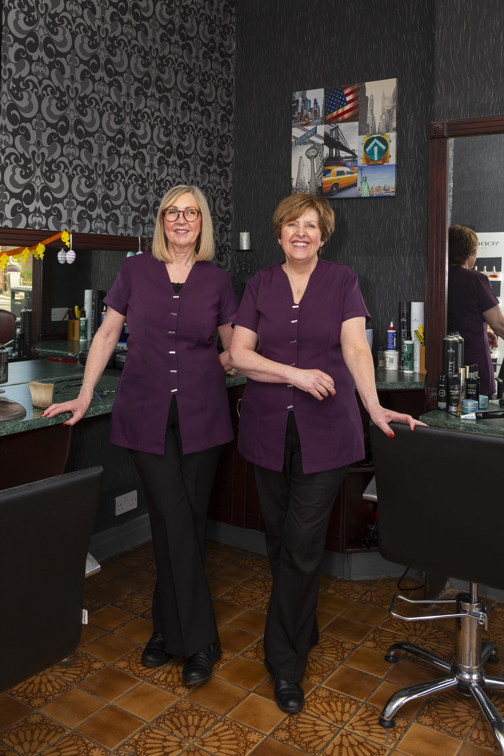 Lynne and Yvonne in Salon 19 – smiling: Lynne and Yvonne, the heart of Salon 19, still smiling after 45+ years of friendship, hairdressing, and community care. Photo by George Ledger