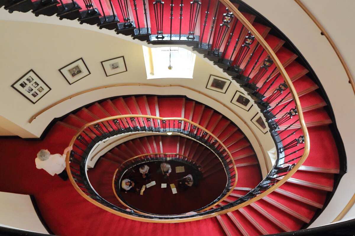 Elliptical red-carpeted staircase spiralling down through the Liverpool Athenaeum with framed pictures on the walls.