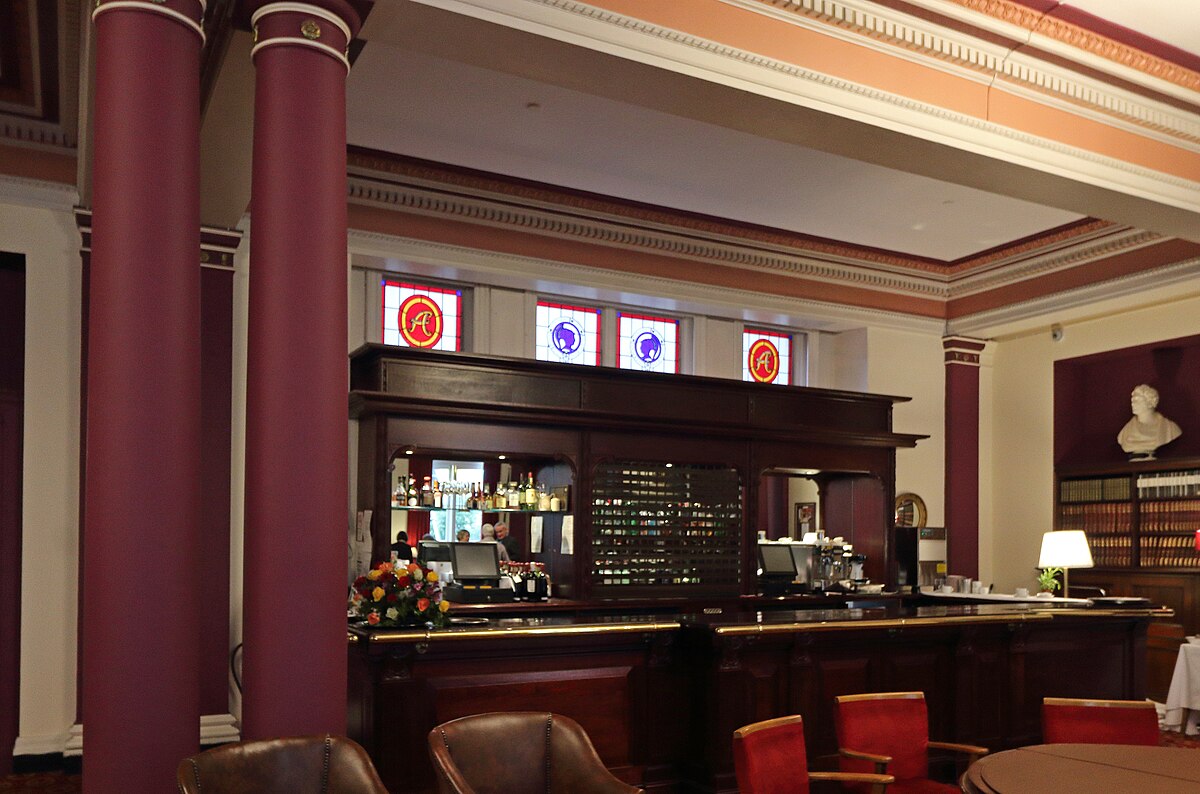 Ornate wooden bar inside the Liverpool Athenaeum newsroom with stained glass windows and classical columns.